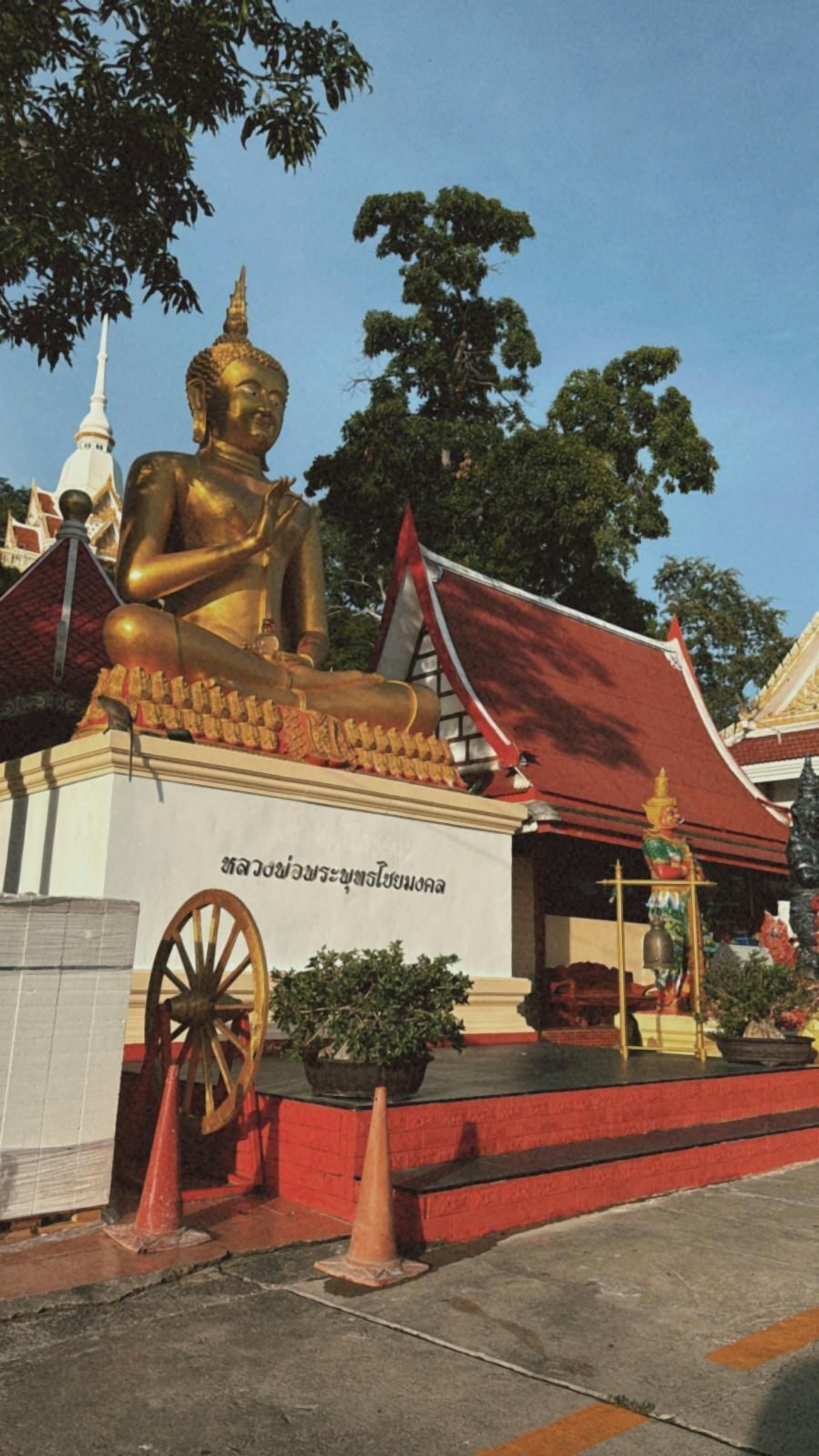 A large golden Buddha statue in front of a temple with a red roof and traditional Thai architecture.