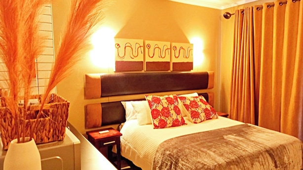 A hotel room with a bed, bedside table, lamp, decorative wall art, and a vase with dried plants.