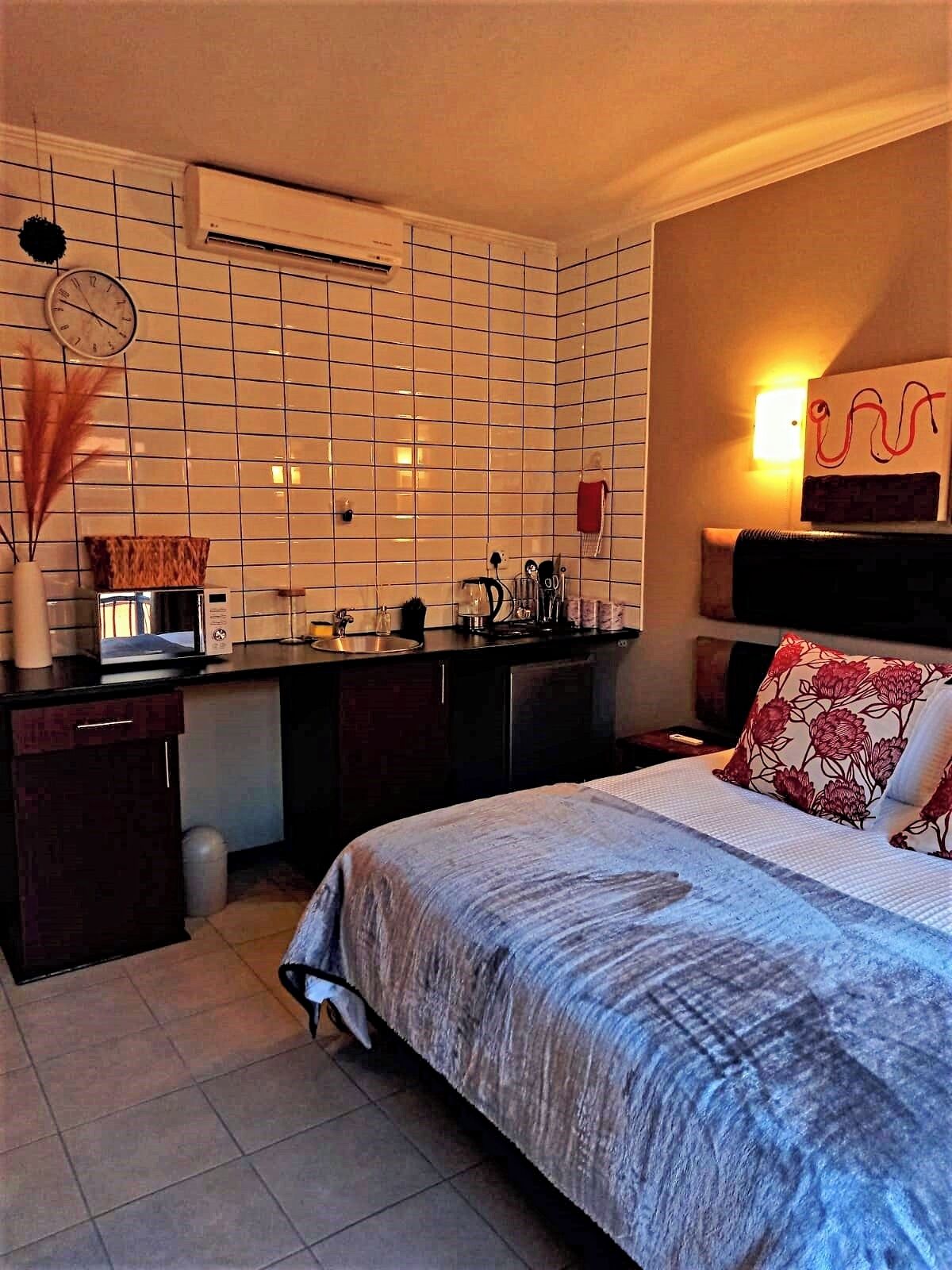A compact hotel room with a kitchenette, a bed with floral pillows, a wall clock, and a decorative wall piece.