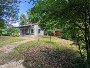 Uma casa com varanda, um pequeno jardim e uma estrutura de madeira ao fundo.