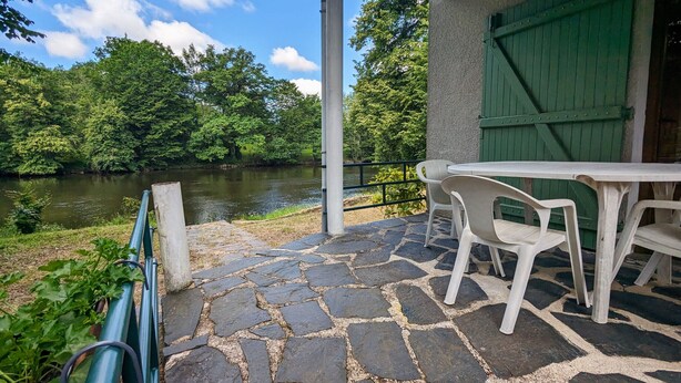 Un patio con suelo de piedra, una mesa y sillas con vistas a un río, con una puerta verde y una barandilla blanca al fondo.