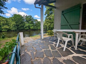 A patio with stone flooring, a table, and chairs overlooking a river, with a green door and a white railing in the background. - Private island in Berry, nature and fishing, central heating (Le Menoux)