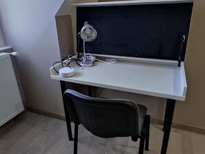 Una taula amb un ordinador, una làmpada i una cadira en una habitació amb un radiador i una paret.