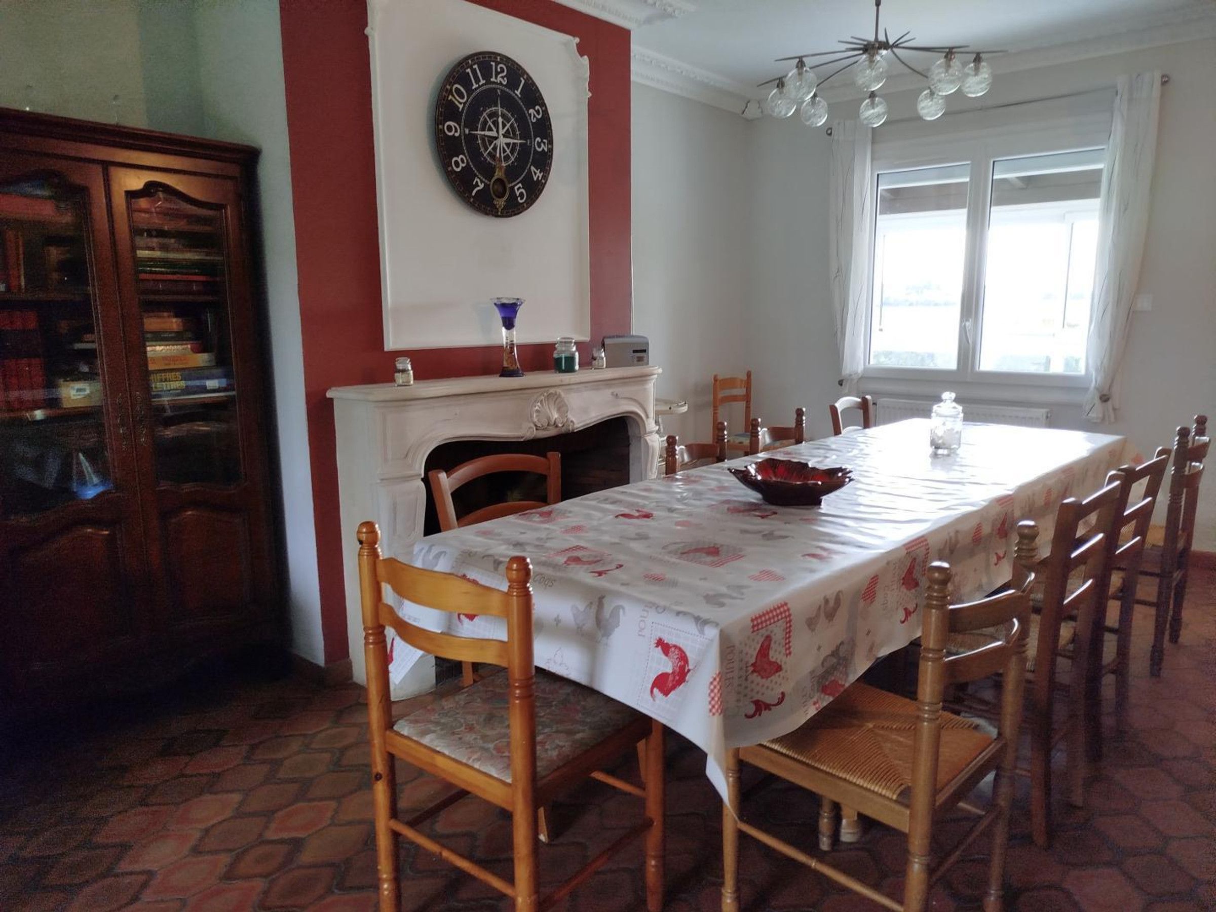 A dining room with a long table, chairs, a fireplace, a clock, and a window.