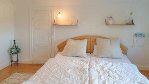 A neatly made bed with white bedding, a wooden headboard, and two bedside tables with lamps. There is a door and a small potted plant on a stand.