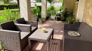 Uma área de pátio ao ar livre com móveis de vime, incluindo dois sofás e uma mesa, rodeada por plantas em vasos e vista para uma área residencial.