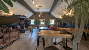 Ruime, overdekte ruimte met houten vloer, een grote tafel en een bar met krukken. Er staan een paar flipperkasten, een grote plant en er is uitzicht naar buiten door ramen.