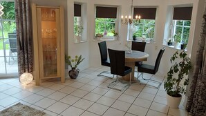 Un comedor con una mesa redonda y sillas, una araña y plantas en macetas junto a las ventanas.