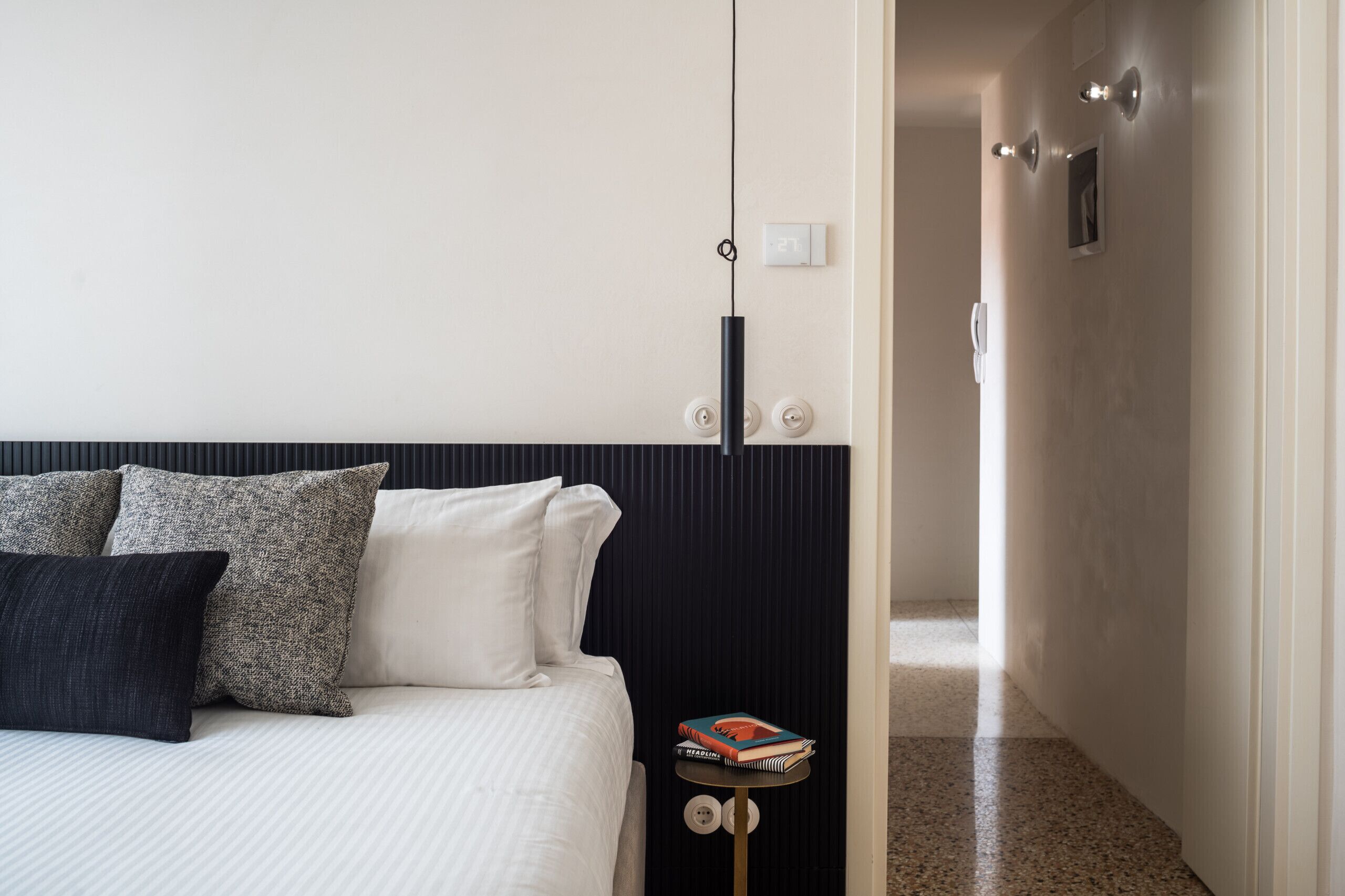A hotel room with a bed, pillows, a bedside table with a book, and a door leading to another room.