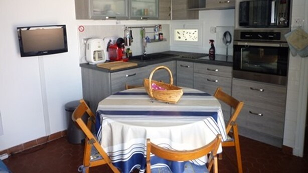 A compact kitchen with a dining area, including a table with chairs, a television mounted on the wall, and modern appliances. - Charming 2-Bed Apartment, A/C, Sleeps 4, Near Beach, Parking Included (Port Leucate)