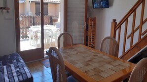 Een kamer met een eettafel, stoelen, een aan de muur bevestigde televisie en een schuifdeur naar een terras met stoelen.
