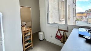 A room with a large window, a wooden shelf with kitchen utensils, a white trash bin, and a desk with a computer and a bag hanging on the wall.