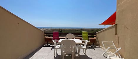 Un balcon donnant sur la plage, avec une table et des chaises, un auvent rouge et une chaise verte.