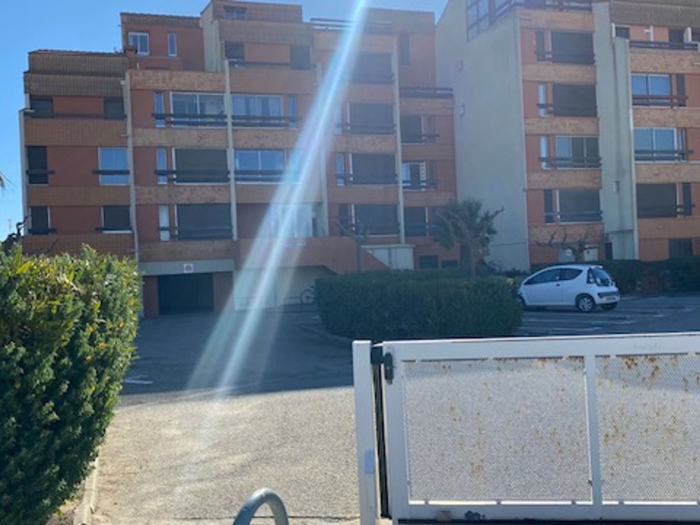 Um edifício residencial de vários andares com varandas, uma área de estacionamento e um carro branco estacionado em frente.