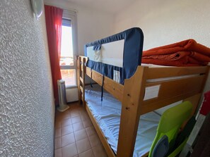 Un lit superposé avec un matelas, un oreiller et une couverture orange. Il y a une fenêtre avec des rideaux rouges et une vue sur l’extérieur.