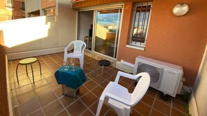 A small outdoor patio with a table covered by a blue cloth, two white plastic chairs, and a white plastic stool. There is a sliding glass door, a window with bars, and an air conditioning unit on the wall. - Charming T2 Cabin, 4 Guests, Near Beach, Terrace, Parking (Port Leucate)