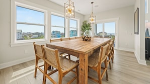 A spacious dining area with a large wooden table and wicker chairs, large windows offering views of the neighborhood, and pendant lighting. - Four Seasons- Endless Ocean and Sound Views, Private Pool, and Hot Tub (Hatteras)
