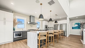 A modern kitchen with white cabinets, a central island with bar stools, stainless steel appliances, and pendant lighting. - Four Seasons- Endless Ocean and Sound Views, Private Pool, and Hot Tub (Hatteras)