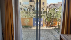 A view through a sliding glass door of a courtyard with a paved ground, a blue fence, and a building in the background. - Charming Garden-Level T2, 4 Guests, Near Beach, Parking Included, Pet-Friendly (Port Leucate)