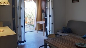 A room with a wooden table, a sofa, and a chair. There is a window with curtains and a view of the outside. - Charming Garden-Level T2, 4 Guests, Near Beach, Parking Included, Pet-Friendly (Port Leucate)