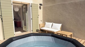 An outdoor hot tub area with a wooden deck, a wooden bench with cushions, and a sliding door leading to a kitchen. - Les Suites du 13 (Aigues-Mortes)