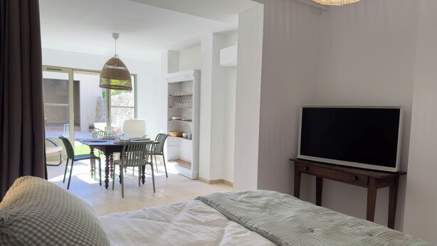 A bedroom with a bed, a TV on a stand, a dining area with chairs and a table, and a view of the outside. - Les Suites du 13 (Aigues-Mortes)