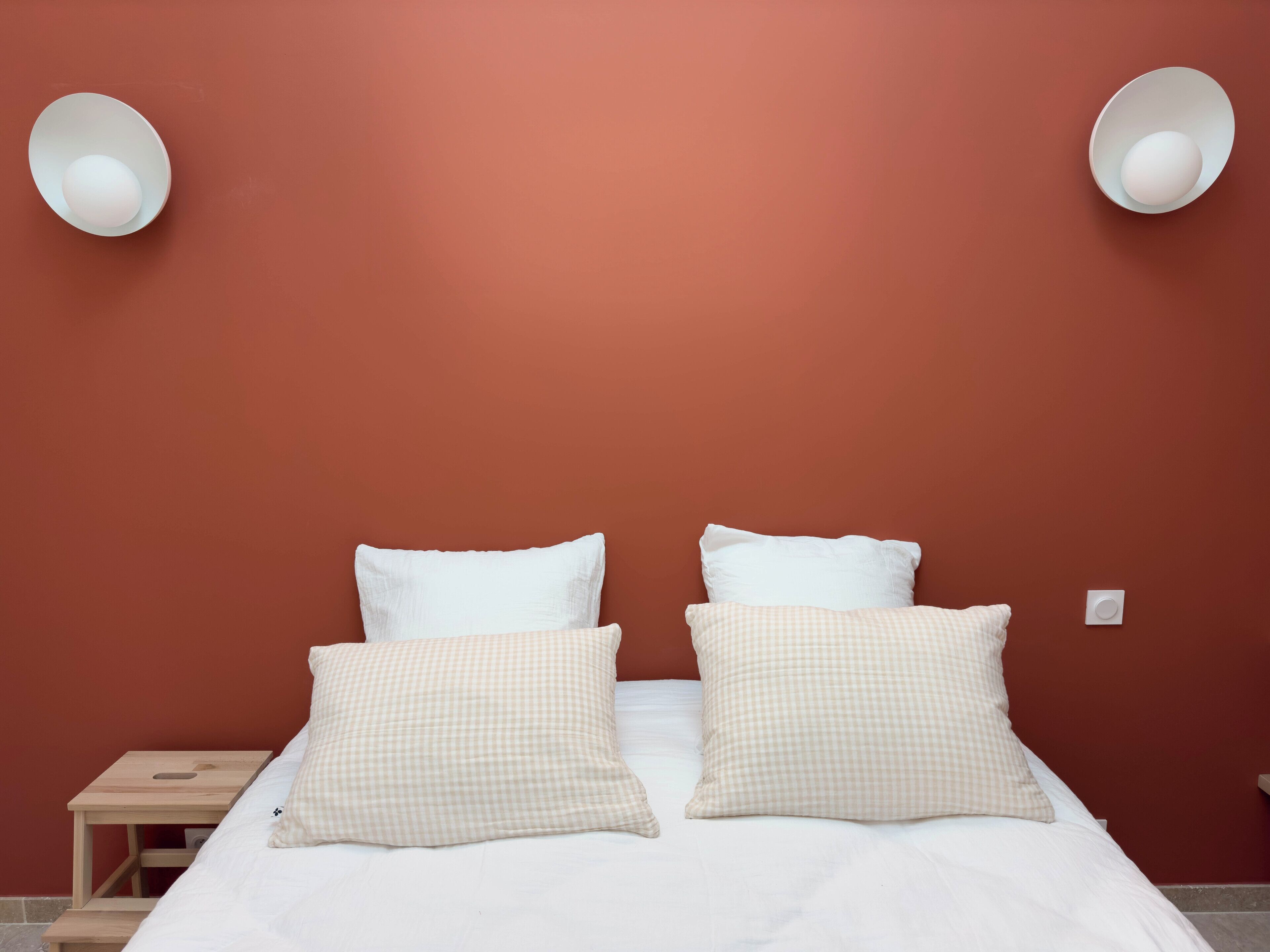 A bed with white bedding and checkered pillows, a wooden nightstand, and a red wall with two wall-mounted lights.