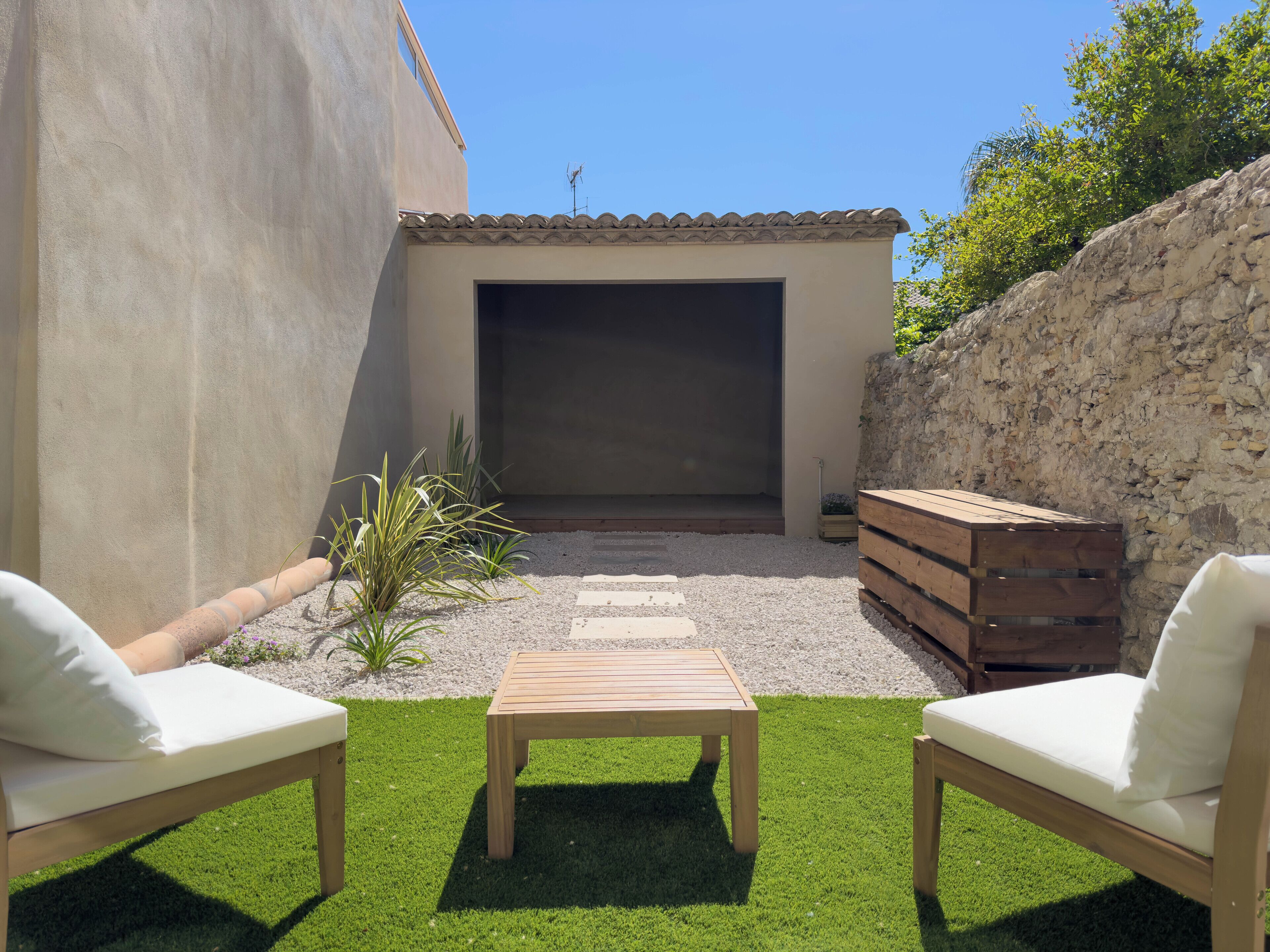 Une terrasse avec deux chaises blanches dotées de coussins, une table en bois et un petit jardin agrémenté de plantes et de gravier.