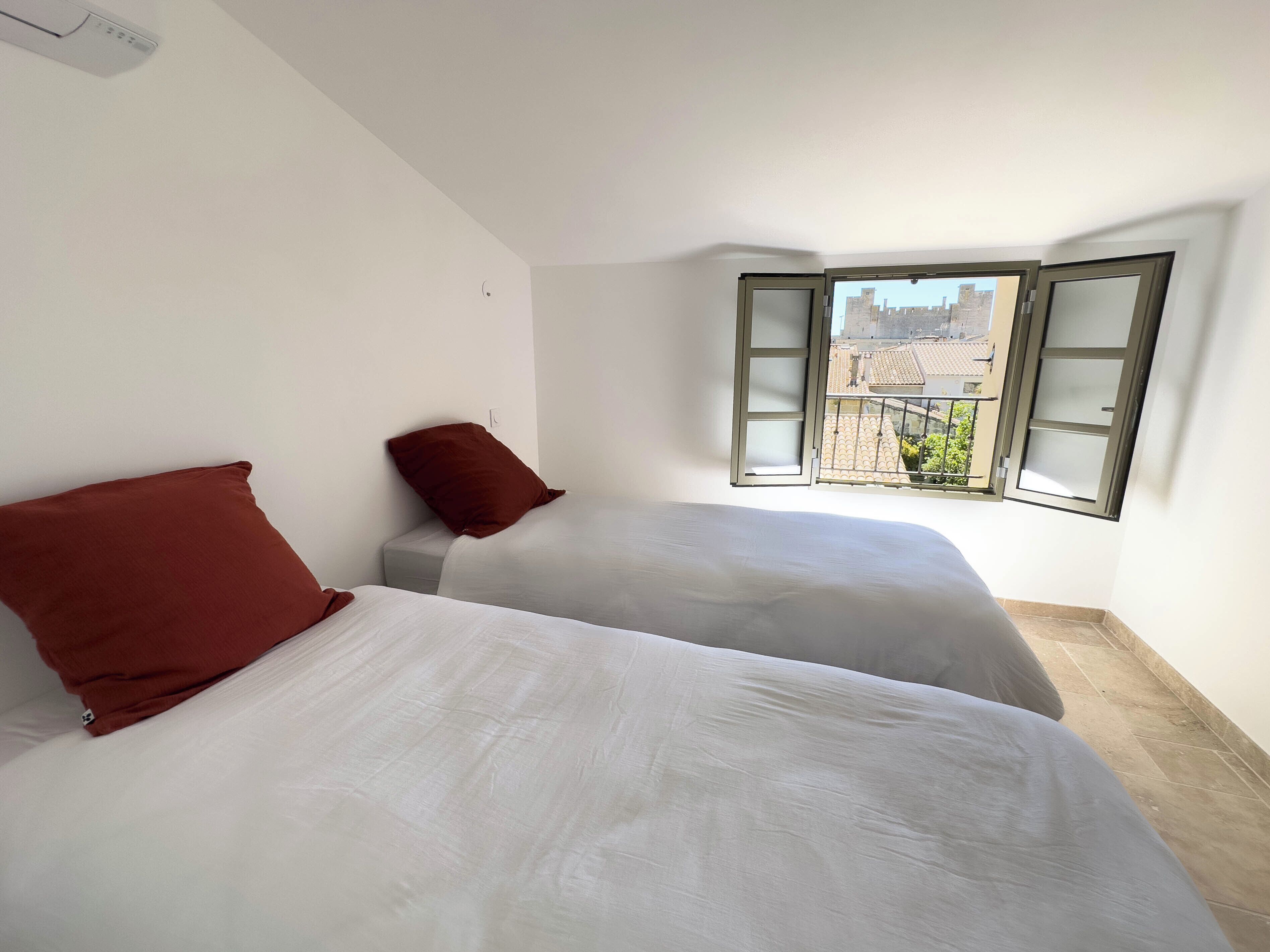 Una habitación con dos camas, una ventana con contraventanas abiertas y vistas a edificios.