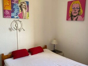 A bedroom with a bed, red pillows, a white duvet, a lamp on a bedside table, and two pop art style paintings on the wall. - Stylish T2 Apartment, 4 Guests, Close to Beach, Safe Parking, Pet-Friendly (Port Leucate)