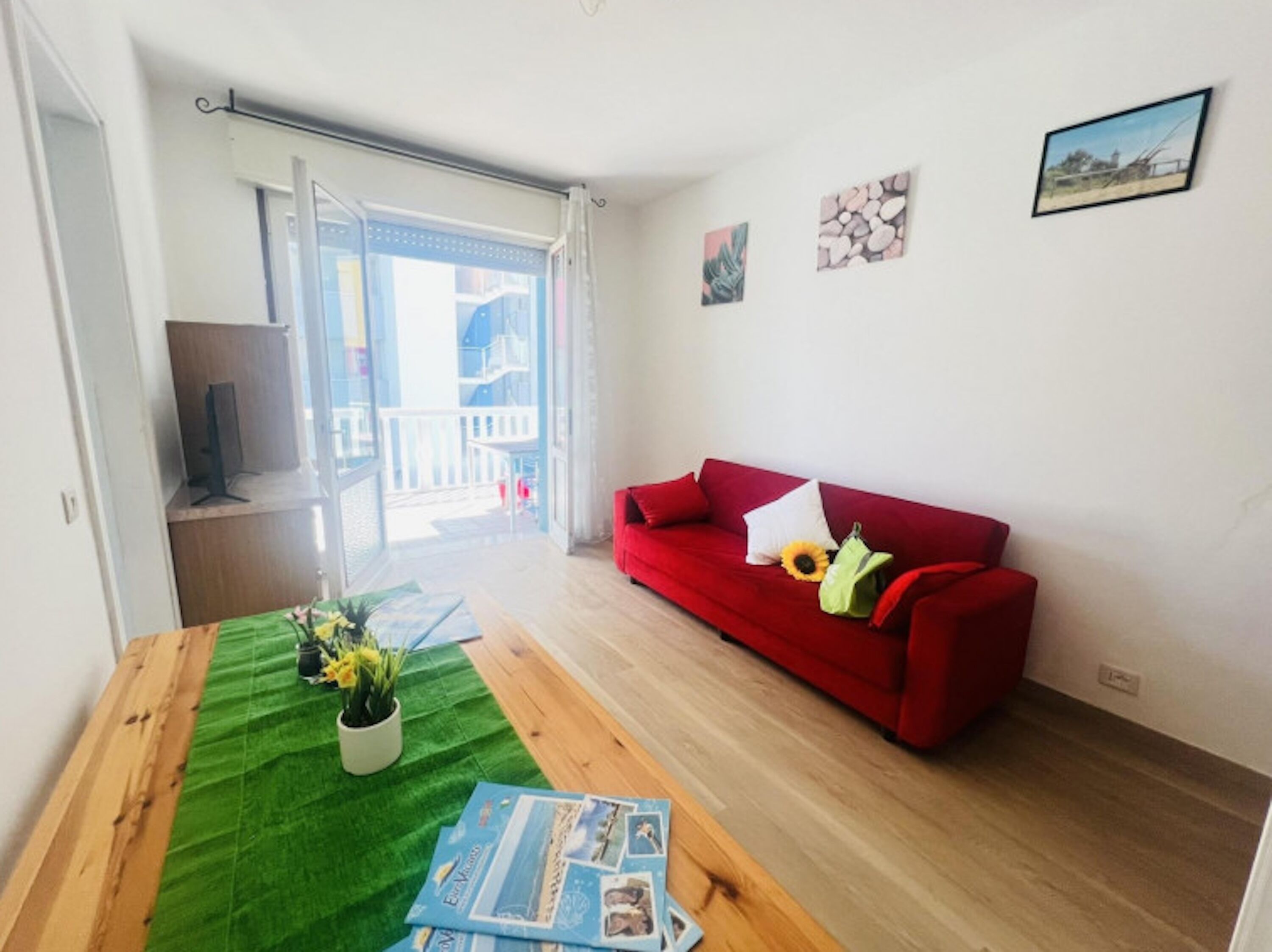 A living room with a red sofa, a wooden table with magazines, a potted plant, and a balcony with a view.