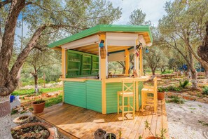 Eine kleine Bar im Freien mit Grün- und Gelbtönen, einer Holzterrasse und ländlicher Umgebung mit Bäumen und Felsen.