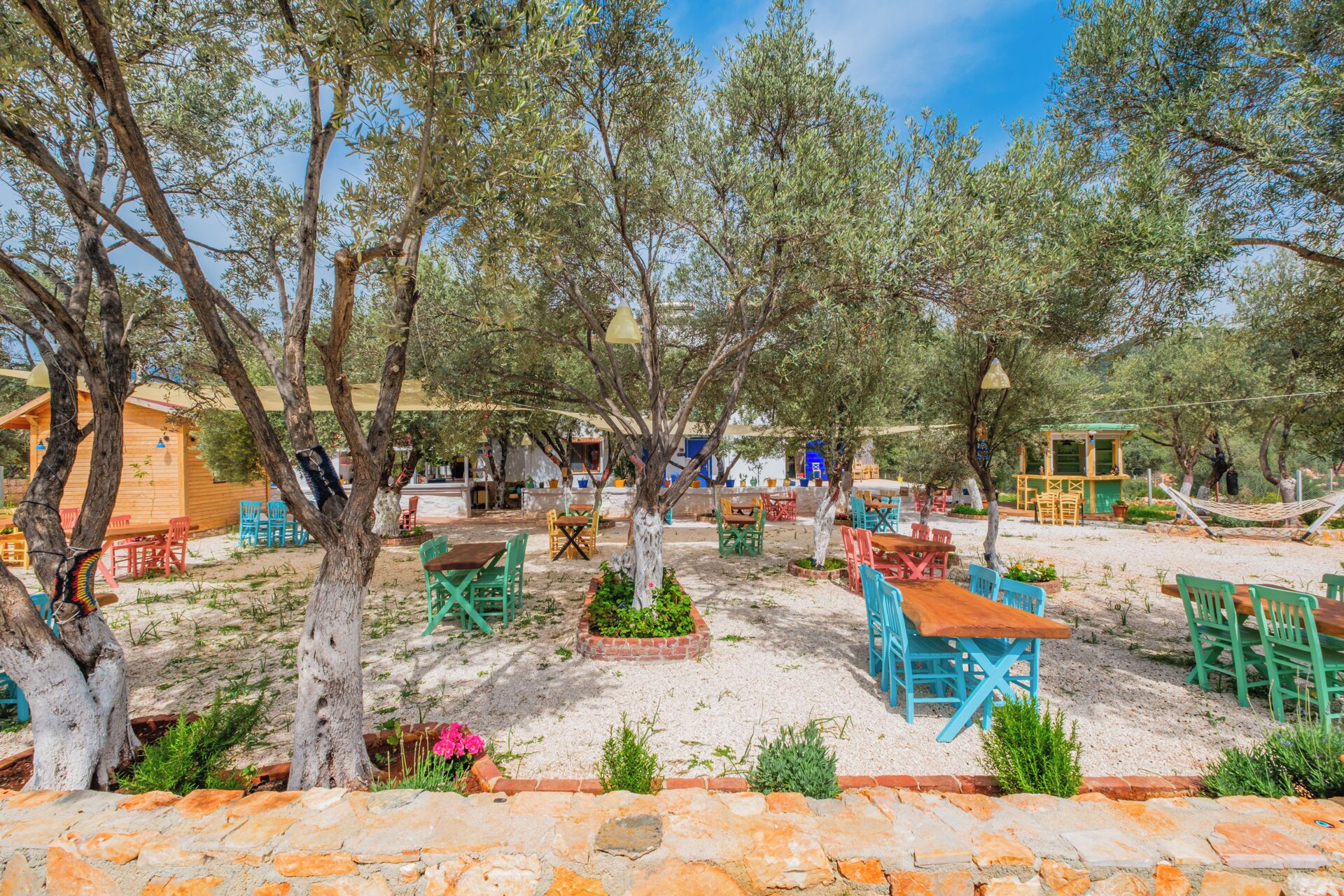 An outdoor seating area with tables and chairs, surrounded by trees and a stone wall.