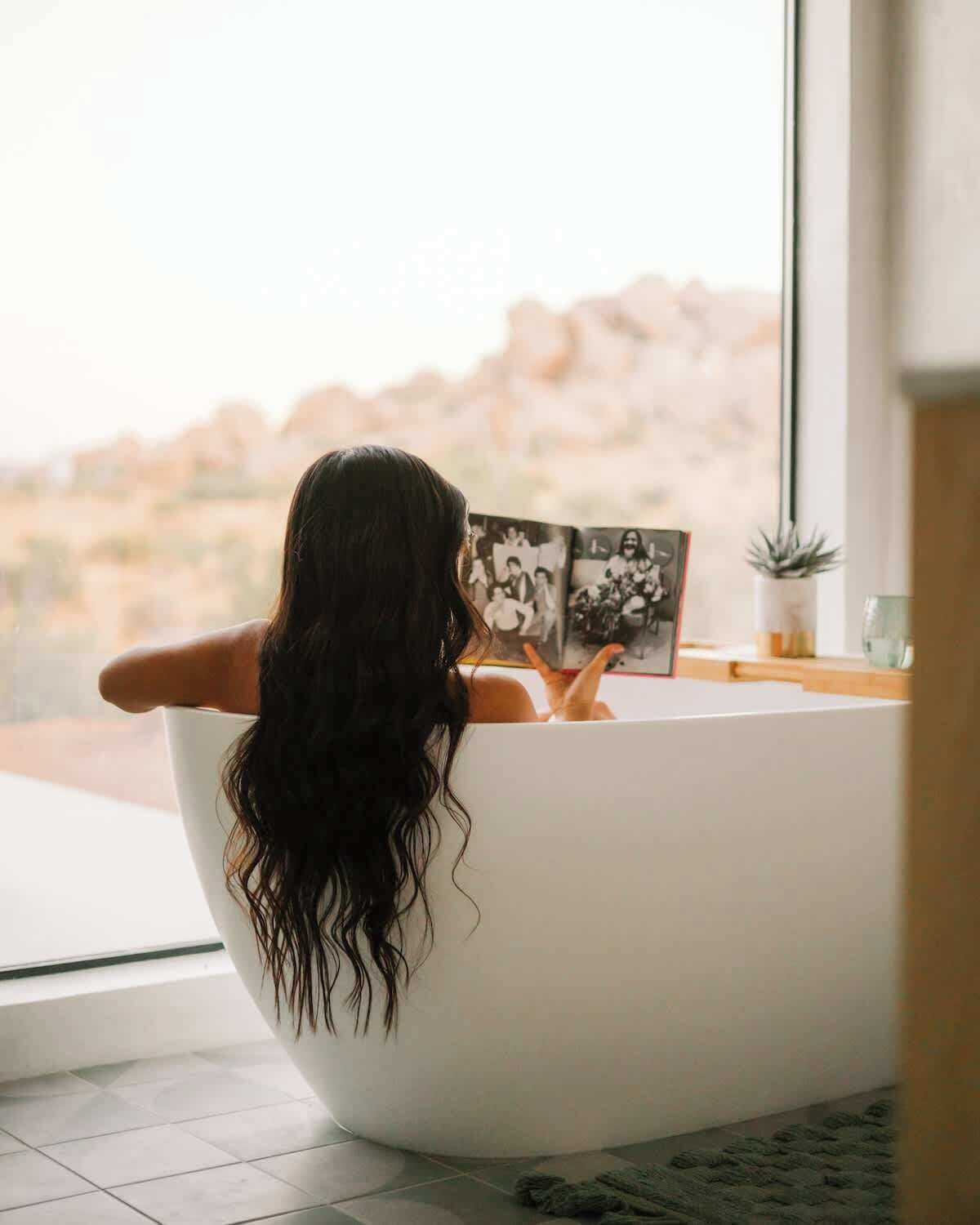 Une personne assise dans une baignoire, avec un livre, une petite plante et un verre à proximité.