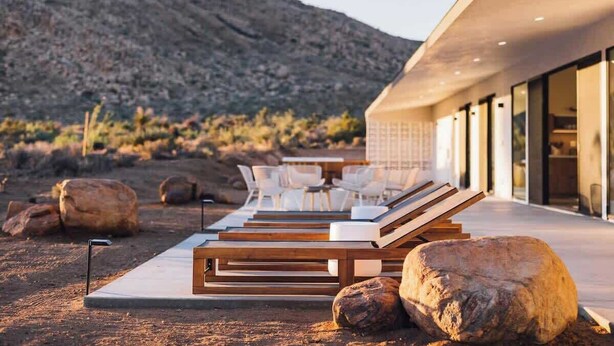 Un moderno espacio lounge al aire libre con tumbonas de madera y sillas blancas, rocas grandes y un paisaje desértico al fondo.