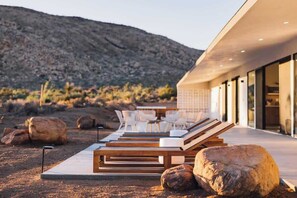 Un espace de détente extérieur moderne, avec des chaises longues en bois et des chaises blanches, de gros rochers et un paysage désertique en arrière-plan.