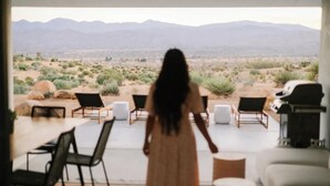 Una mujer vestida caminando por una habitación con vistas al desierto, un área de comedor y asientos.