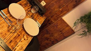 Una mesa de madera para comer con platos blancos, cubiertos y una copa de vino. Se ve un reloj de pared y una lámpara colgante.