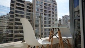 Un balcón con dos sillas blancas y una mesa de vidrio, con vista a un edificio de gran altura.