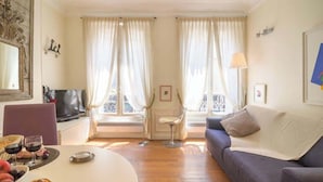 A living room with a sofa, a TV, a dining table with food, and two windows with curtains. - Le Marais Moderne (Paris)