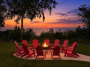 Ein kreisförmiges Arrangement aus roten Adirondack-Stühlen um eine Feuerstelle mit Blick auf den Ozean bei Sonnenuntergang.
