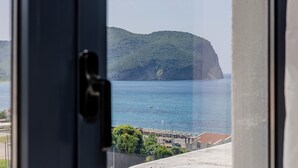 A coastal view through a window, featuring a mountainous landscape, a body of water, and a parking area with buildings. - Hotel Aquilo (Petrovac)