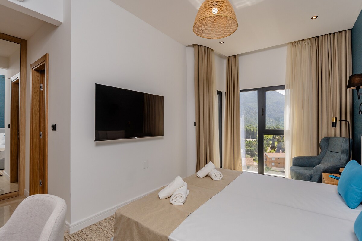 A modern hotel room with a large bed, a flat-screen TV, a seating area with a sofa and armchairs, and a view of the outdoors.