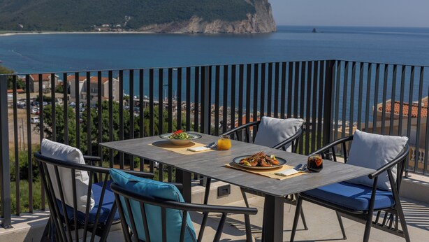 A balcony with a table set for two, overlooking a coastal landscape with a mountain in the distance. - Hotel Aquilo (Petrovac)