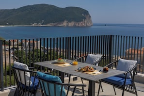 A balcony with a table set for two, overlooking a coastal landscape with a mountain in the distance. - Hotel Aquilo (Petrovac)