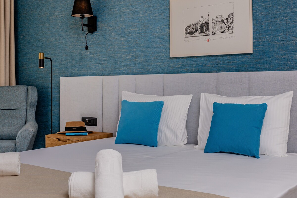A neatly made bed with white and blue pillows, a grey upholstered armchair, and a bedside table with a hat and a book.