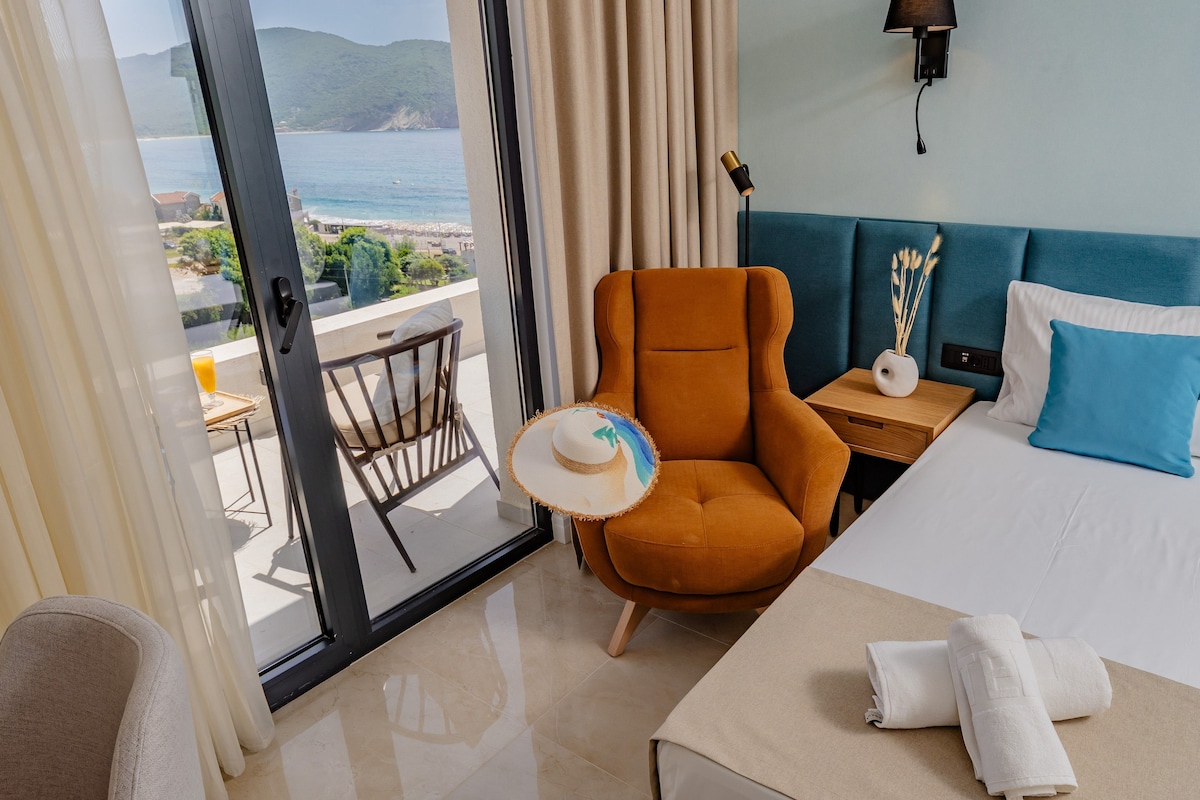 A hotel room with a bed, a chair, a small table, and a view of the sea.