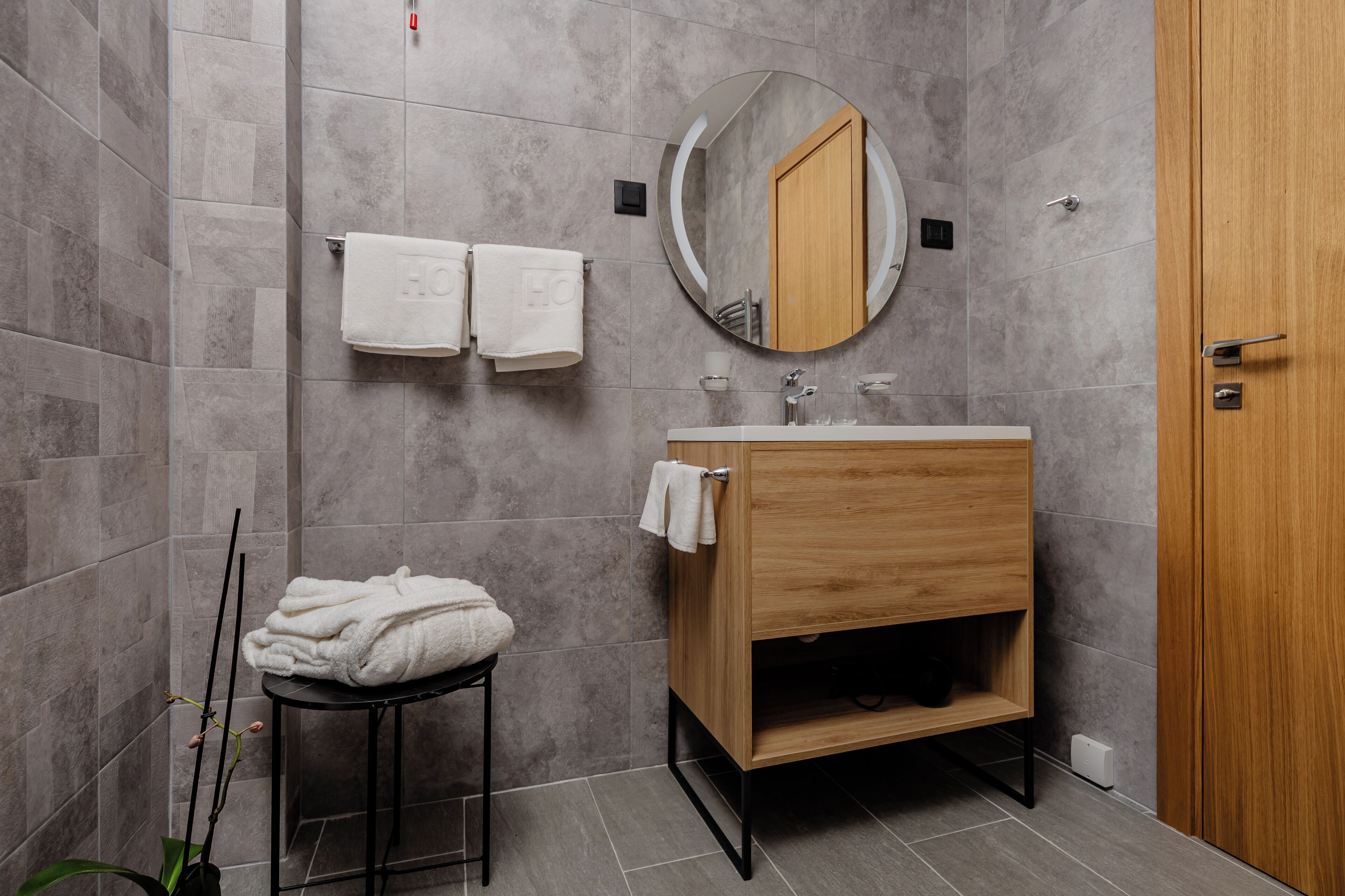 A modern bathroom with grey tiles, a wooden vanity, a round mirror, and two towels hanging on hooks.