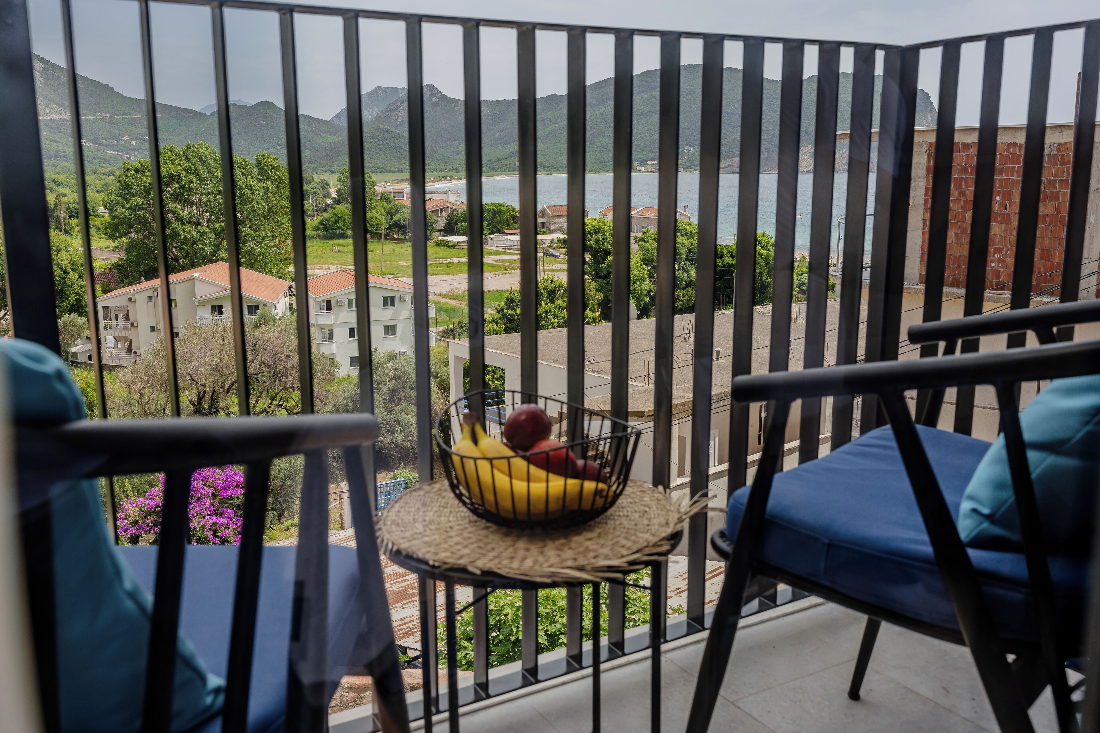 A balcony with a view of a mountainous landscape and a body of water. There is a small table with a fruit basket on it.