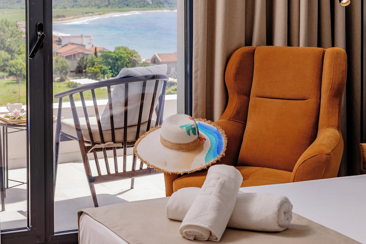 A hotel room with a large window, a view of the beach and mountains, a brown armchair, a white bed, and a straw hat on the chair.